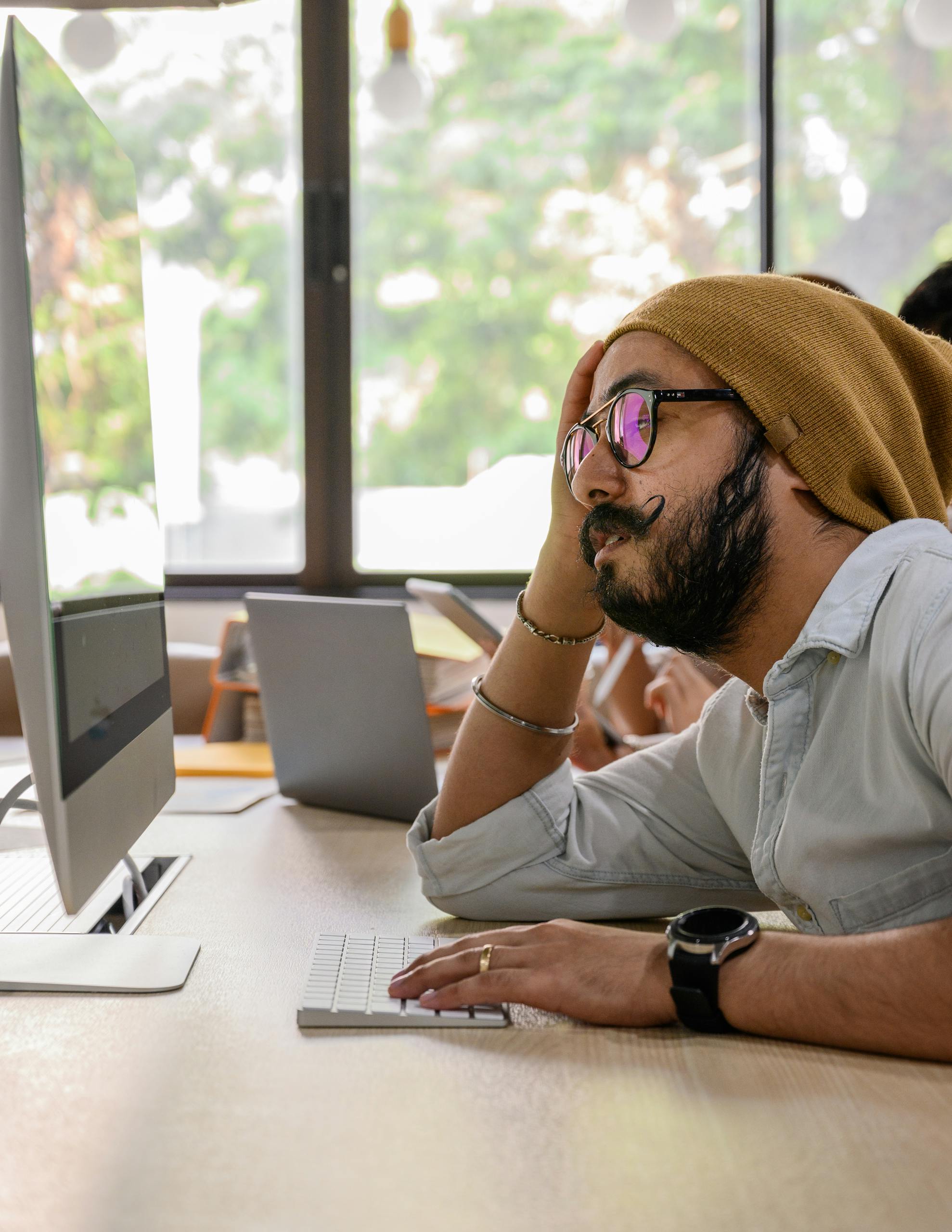 A man with glasses and hat working on a computer in a modern office with natural light.